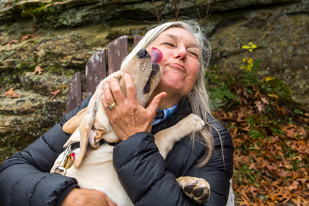 A dog sitting in an older woman's lap and licking her face.